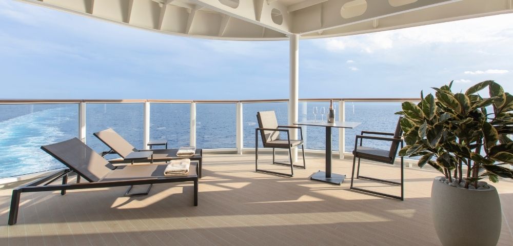 Spacious cruise ship deck with modern loungers and chairs, a small table, and a potted plant. Ocean views and clear blue sky create a serene atmosphere.