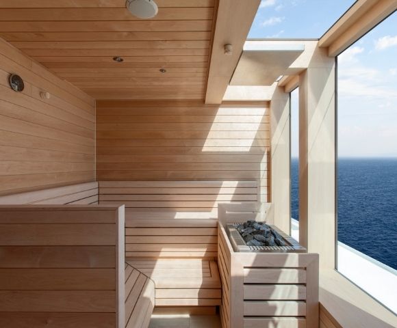 A modern wooden sauna with large windows overlooking the ocean. Sunlight filters in, creating a serene and relaxing atmosphere.