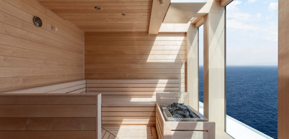 A modern wooden sauna with large windows overlooking the ocean. Sunlight filters in, creating a serene and relaxing atmosphere.