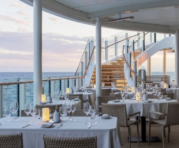 Elegant outdoor dining area on a ship deck at sunset, featuring white tablecloths, wine glasses, and soft lanterns. A staircase leads to an upper level.