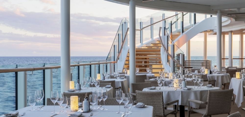 Elegant outdoor dining area on a ship deck at sunset, featuring white tablecloths, wine glasses, and soft lanterns. A staircase leads to an upper level.