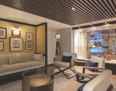 Elegant living room with a beige sofa, modern chairs, glass-topped table, framed art, and a ceiling spotlight. Decor includes nautical-themed pieces.