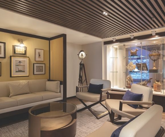 Elegant living room with a beige sofa, modern chairs, glass-topped table, framed art, and a ceiling spotlight. Decor includes nautical-themed pieces.