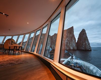 Luxurious cruise ship interior with large curved windows showcasing a breathtaking view of rocky sea cliffs under a moody, overcast sky.