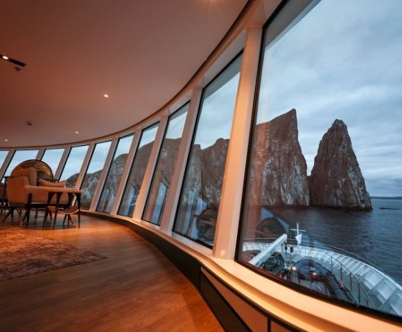 Luxurious cruise ship interior with large curved windows showcasing a breathtaking view of rocky sea cliffs under a moody, overcast sky.