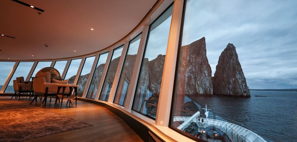 Luxurious cruise ship interior with large curved windows showcasing a breathtaking view of rocky sea cliffs under a moody, overcast sky.