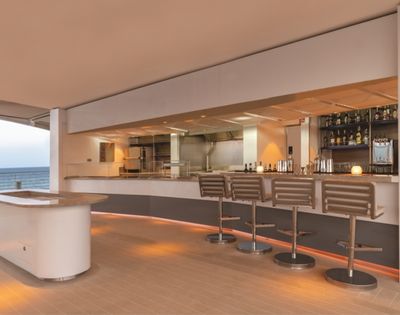 Sleek modern bar with high stools and a well-stocked shelf, overlooking the ocean. Warm lighting creates a relaxed, upscale atmosphere.