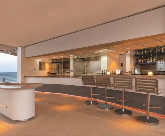 Sleek modern bar with high stools and a well-stocked shelf, overlooking the ocean. Warm lighting creates a relaxed, upscale atmosphere.