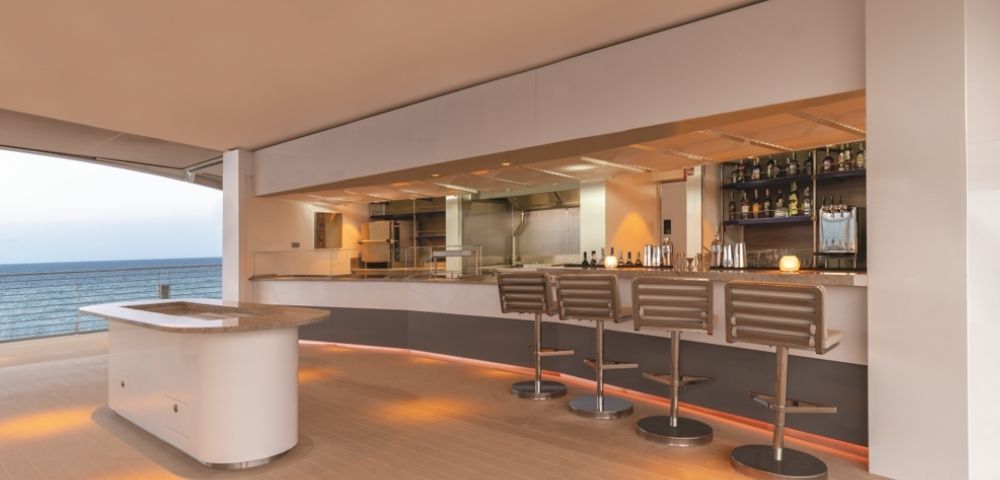 Sleek modern bar with high stools and a well-stocked shelf, overlooking the ocean. Warm lighting creates a relaxed, upscale atmosphere.