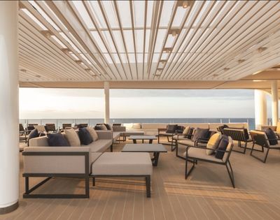 Spacious deck with modern couches and chairs under a slatted roof, overlooking a calm ocean at sunset, creating a serene and relaxed atmosphere.