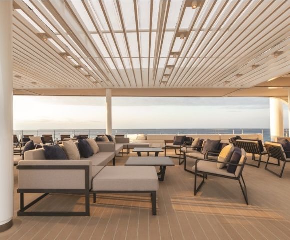 Spacious deck with modern couches and chairs under a slatted roof, overlooking a calm ocean at sunset, creating a serene and relaxed atmosphere.