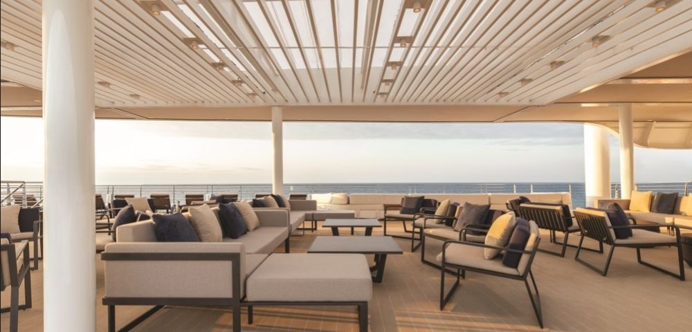 Spacious deck with modern couches and chairs under a slatted roof, overlooking a calm ocean at sunset, creating a serene and relaxed atmosphere.