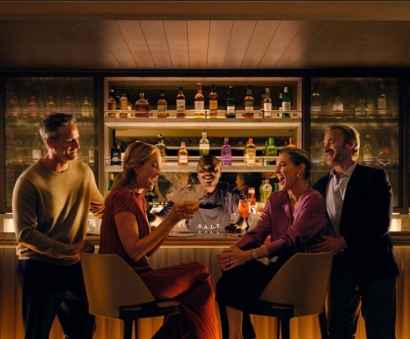 A group of four people, two men and two women, laughing and talking at a stylish, warmly lit bar with a bartender and shelves of liquor bottles behind them.