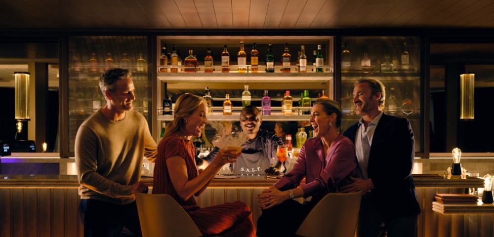 A group of four people, two men and two women, laughing and talking at a stylish, warmly lit bar with a bartender and shelves of liquor bottles behind them.