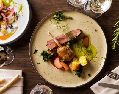Aerial view of a gourmet dish featuring lamb, green puree, vegetables, and herbs on a beige plate. Nearby, a seafood platter and glasses of white wine.