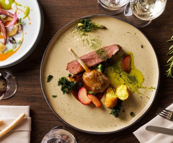 Aerial view of a gourmet dish featuring lamb, green puree, vegetables, and herbs on a beige plate. Nearby, a seafood platter and glasses of white wine.