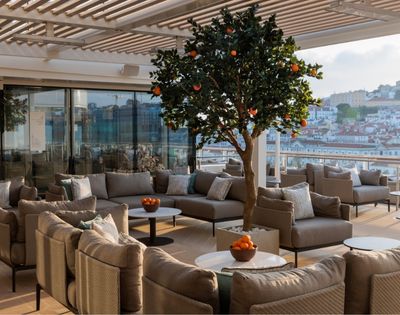 Rooftop terrace with modern beige seating, a central orange tree, and bowls of fruit. Glass doors and a scenic city view create a serene ambiance.