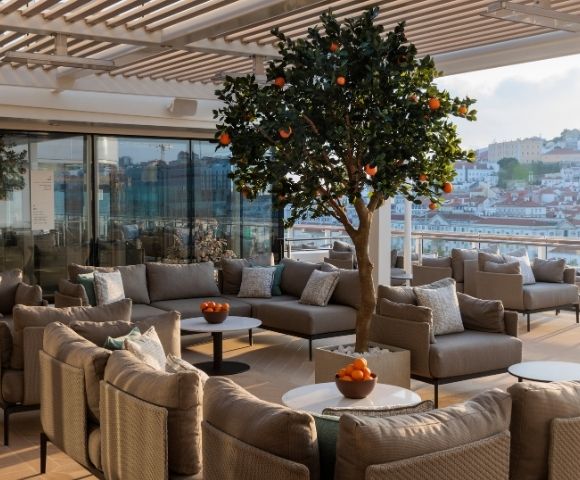 Rooftop terrace with modern beige seating, a central orange tree, and bowls of fruit. Glass doors and a scenic city view create a serene ambiance.