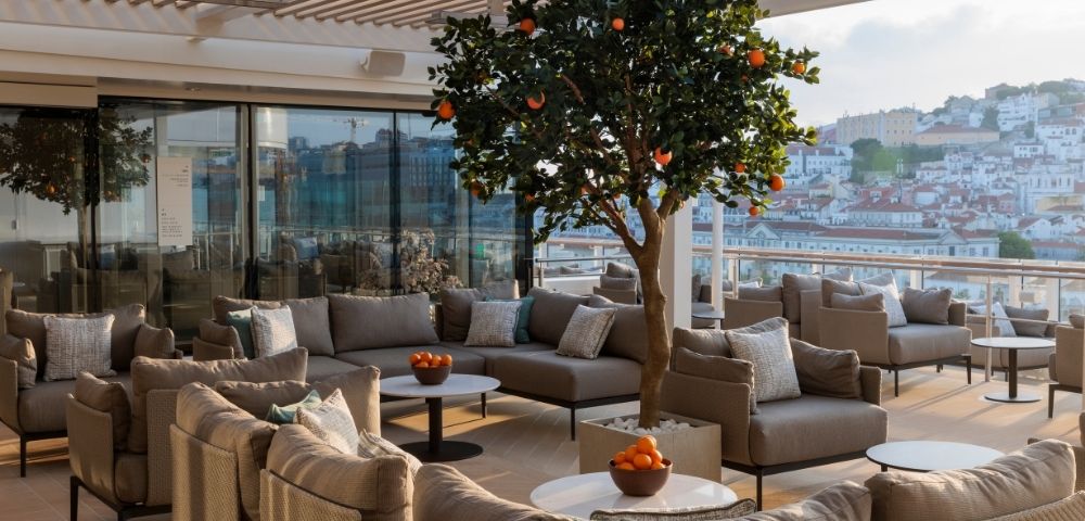 Rooftop terrace with modern beige seating, a central orange tree, and bowls of fruit. Glass doors and a scenic city view create a serene ambiance.