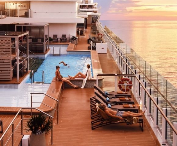 Sunset view from a cruise ship's deck with a pool. People relaxing on loungers, enjoying the serene sea and vibrant sky, creating a tranquil atmosphere.