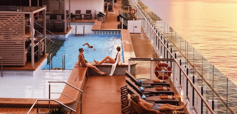 Sunset view from a cruise ship's deck with a pool. People relaxing on loungers, enjoying the serene sea and vibrant sky, creating a tranquil atmosphere.