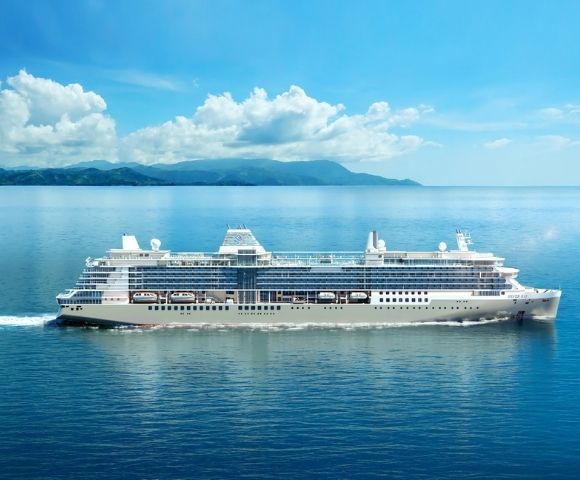 A large, white cruise ship sails on clear blue sea under a bright sky with fluffy clouds. Distant green hills loom on the horizon, conveying a tranquil scene.