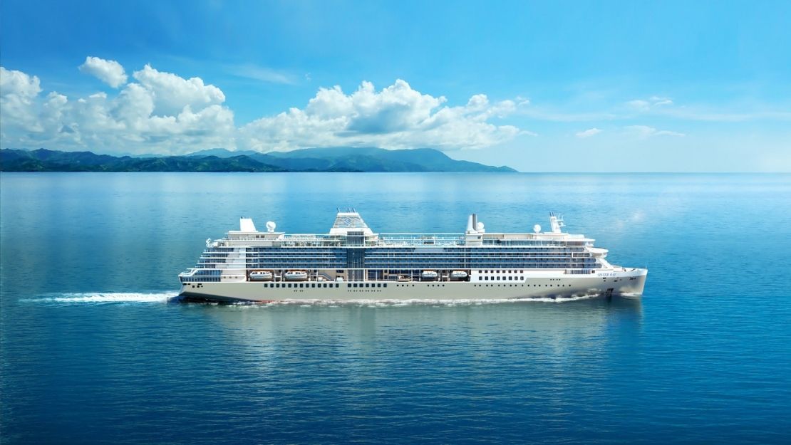 A large, white cruise ship sails on clear blue sea under a bright sky with fluffy clouds. Distant green hills loom on the horizon, conveying a tranquil scene.