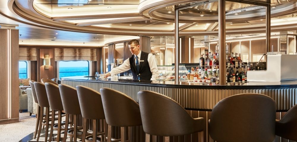 Luxurious cruise ship bar with elegant chairs and a polished counter. A bartender adjusts glassware, conveying a relaxed, upscale atmosphere.