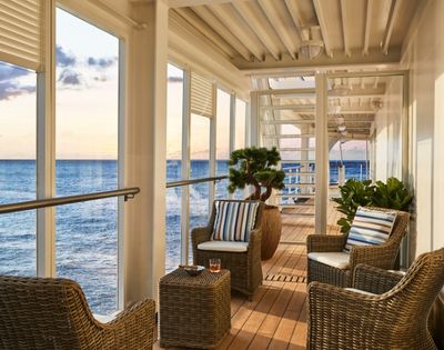 Cozy balcony overlooking the ocean at sunset, featuring wicker chairs with striped cushions, a small table, potted plants, and a serene ambiance.