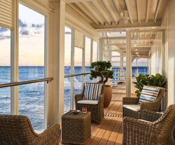 Cozy balcony overlooking the ocean at sunset, featuring wicker chairs with striped cushions, a small table, potted plants, and a serene ambiance.