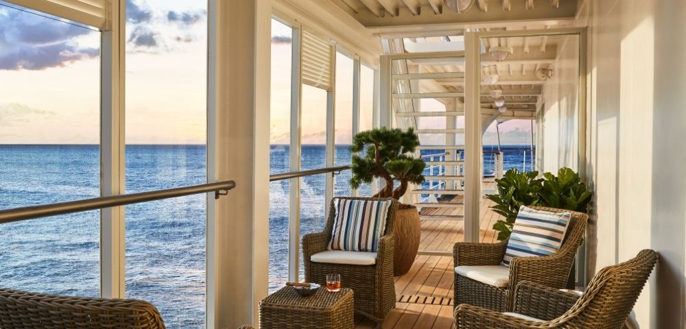 Cozy balcony overlooking the ocean at sunset, featuring wicker chairs with striped cushions, a small table, potted plants, and a serene ambiance.