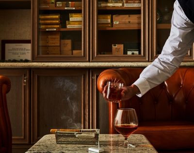A person serves a drink in a cozy, upscale cigar lounge with brown leather couches. A cigar rests in an ashtray, with smoke gently curling upwards.