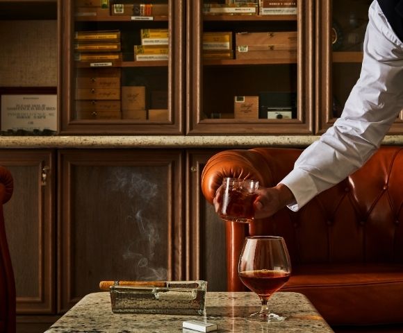 A person serves a drink in a cozy, upscale cigar lounge with brown leather couches. A cigar rests in an ashtray, with smoke gently curling upwards.