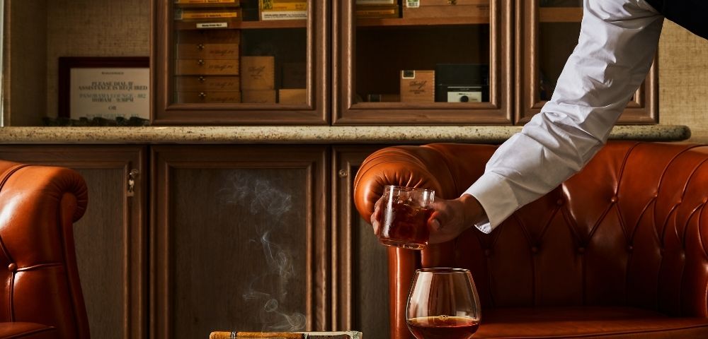 A person serves a drink in a cozy, upscale cigar lounge with brown leather couches. A cigar rests in an ashtray, with smoke gently curling upwards.