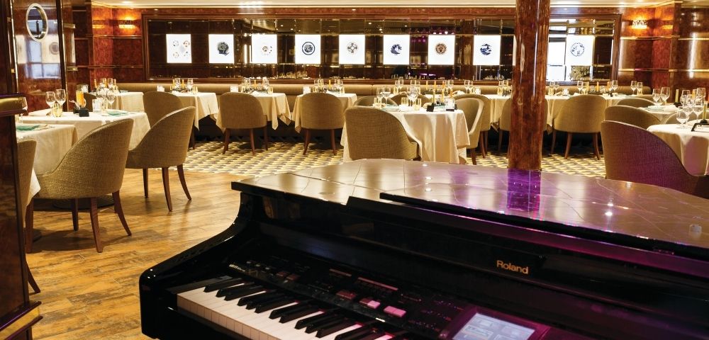 Grand piano in an elegant restaurant with round tables and beige chairs. Soft lighting and wall art create a sophisticated and inviting atmosphere.