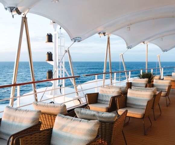 Spacious ship deck with wicker chairs and striped cushions, under a white canopy. Calm ocean and clear sky create a peaceful and luxurious atmosphere.