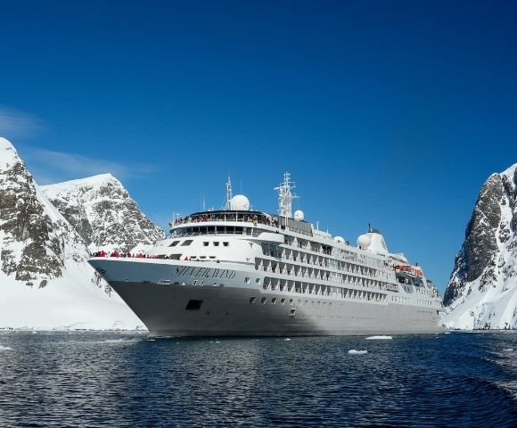 A large white cruise ship sails between snow-covered mountains under a clear blue sky, evoking a sense of adventure and exploration.