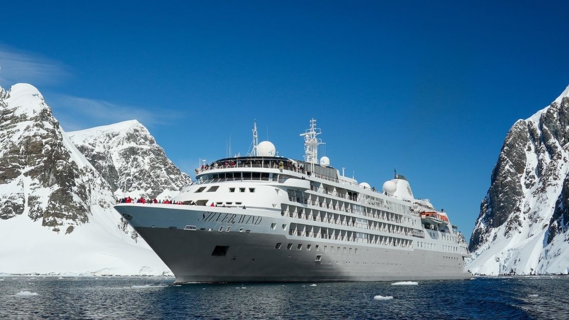 A large white cruise ship sails between snow-covered mountains under a clear blue sky, evoking a sense of adventure and exploration.