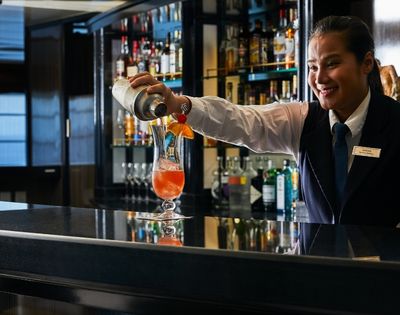 A bartender in uniform smiles while pouring a cocktail into a glass on a bar counter. The bar is well-stocked, conveying a warm, inviting atmosphere.