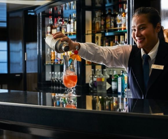 A bartender in uniform smiles while pouring a cocktail into a glass on a bar counter. The bar is well-stocked, conveying a warm, inviting atmosphere.