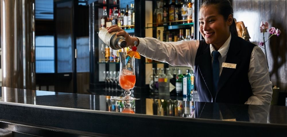 A bartender in uniform smiles while pouring a cocktail into a glass on a bar counter. The bar is well-stocked, conveying a warm, inviting atmosphere.