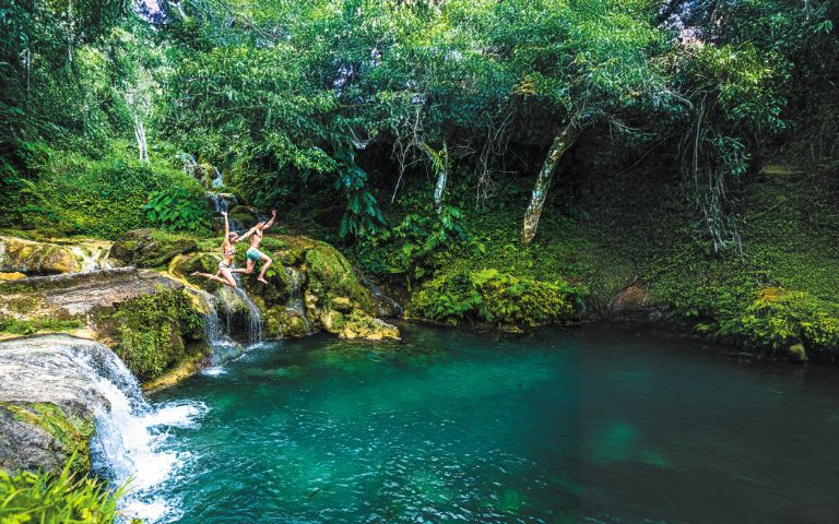 Two people joyfully jump into a clear, turquoise pool surrounded by lush greenery and cascading waterfalls in a tranquil natural setting.
