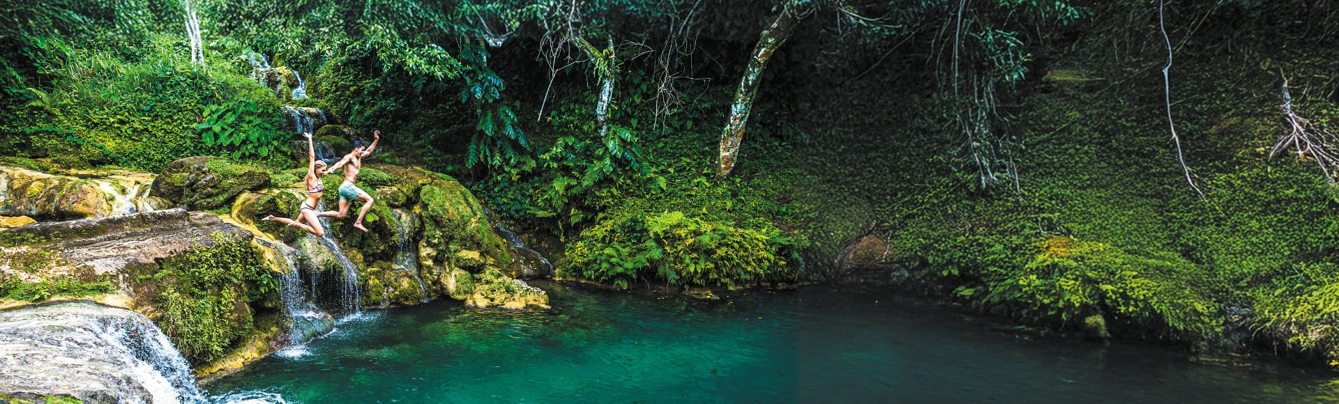 Two people joyfully jump into a clear, turquoise pool surrounded by lush greenery and cascading waterfalls in a tranquil natural setting.