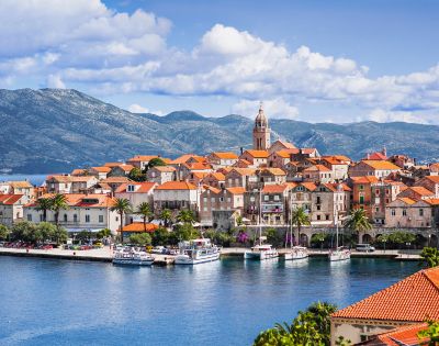 Coastal town with terracotta-roofed buildings and a prominent church tower, set against a backdrop of mountains and a cloudy blue sky, evokes tranquility.