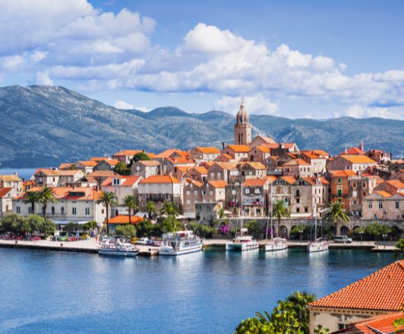 Coastal town with terracotta-roofed buildings and a prominent church tower, set against a backdrop of mountains and a cloudy blue sky, evokes tranquility.