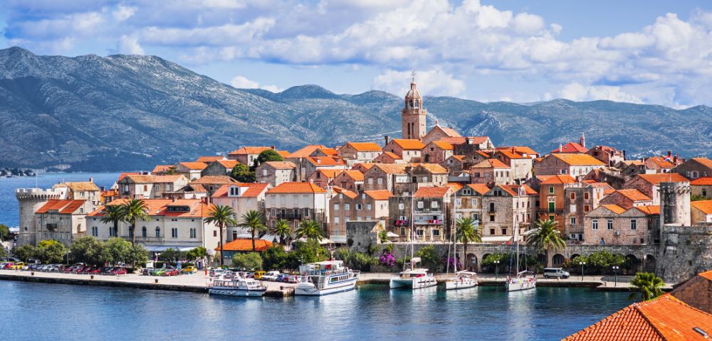 Coastal town with terracotta-roofed buildings and a prominent church tower, set against a backdrop of mountains and a cloudy blue sky, evokes tranquility.