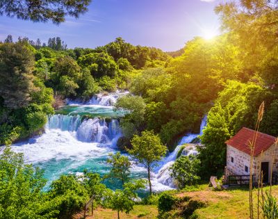Sunlit landscape featuring cascading waterfalls surrounded by lush green trees, with a small stone building and red roof in the foreground.