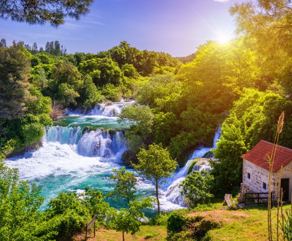 Sunlit landscape featuring cascading waterfalls surrounded by lush green trees, with a small stone building and red roof in the foreground.