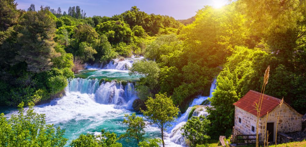 Sunlit landscape featuring cascading waterfalls surrounded by lush green trees, with a small stone building and red roof in the foreground.