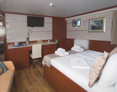 A cozy ship cabin features a neatly made bed with folded towels, a desk with a chair, a wall-mounted TV, and a small window, creating a warm, inviting atmosphere.
